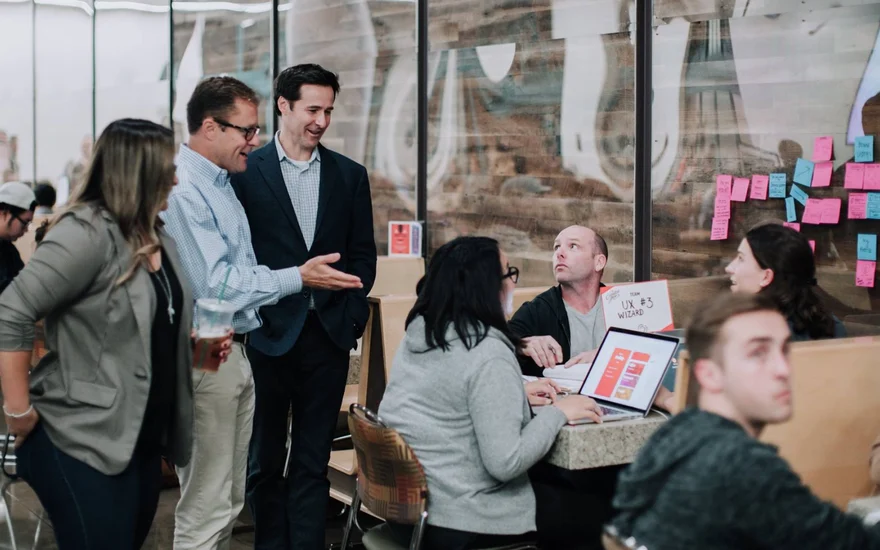 Judges and mentors stop by the team table, with the sticky-note flow on the wall behind them.
